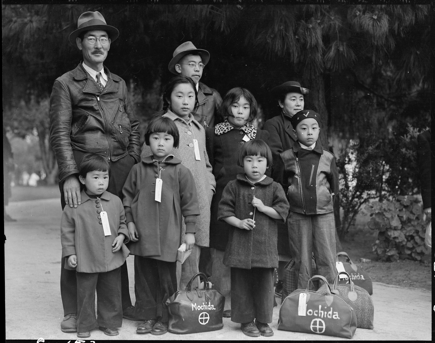 Original caption: Hayward, California. Members of the Mochida family awaiting evacuation bus. Identification tags are used to aid in keeping the family unit intact during all phases of evacuation. Mochida operated a nursery and five greenhouses on a two-acre site in Eden Township. He raised snapdragons and sweet peas. Evacuees of Japanese ancestry will be housed in War Relocation Authority centers for the duration.