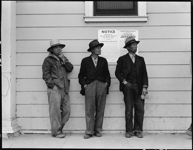 Byron, California. Field laborers of Japanese ancestry in front of Wartime Civil Control Administration station where they have come for instructions and assistance in regard to their evacuation due in three days under Civilian Exclusion Order Number 24. This order affects 850 persons in this area. The men are now waiting for the truck which will take them, with the rest of the field crew, back to the large-scale delta ranch.